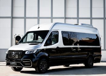 A black and white Mercedes-Benz Sprinter van, a unique creation, is parked outside in front of a large, gray industrial building that resembles a garage.
