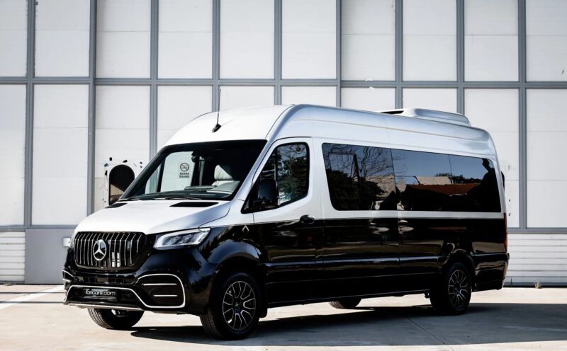 A black and white Mercedes-Benz Sprinter van, a unique creation, is parked outside in front of a large, gray industrial building that resembles a garage.