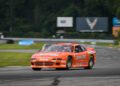 An orange Dodge race car with "Earnhardt" branding races at Laguna Seca during the Rolex Monterey Motorsports Reunion, surrounded by greenery and advertising boards, as part of the IROC All-Star Legends event.