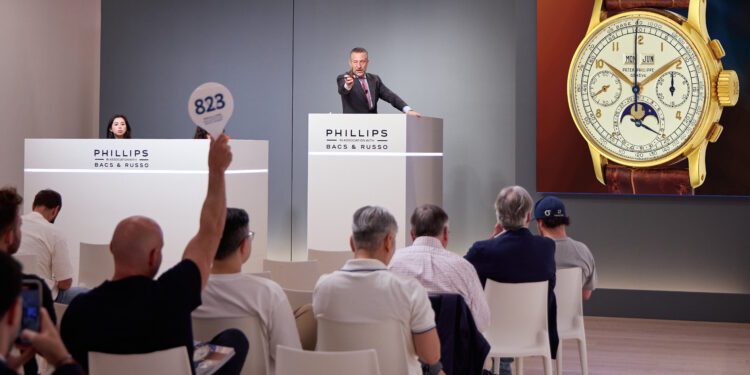 At a New York Watch Auction hosted by Phillips, the auctioneer takes bids on a luxury wristwatch as a participant raises a numbered paddle and others watch the proceedings.