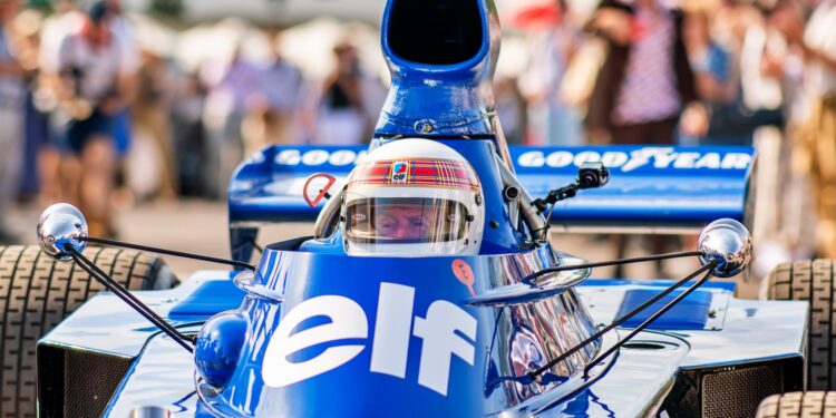 A driver in a blue vintage Formula 1 car with "elf" and "Goodyear" logos prepares to race at the Goodwood Festival, surrounded by a crowd, showcasing one of the era-defining cars of motorsport history.