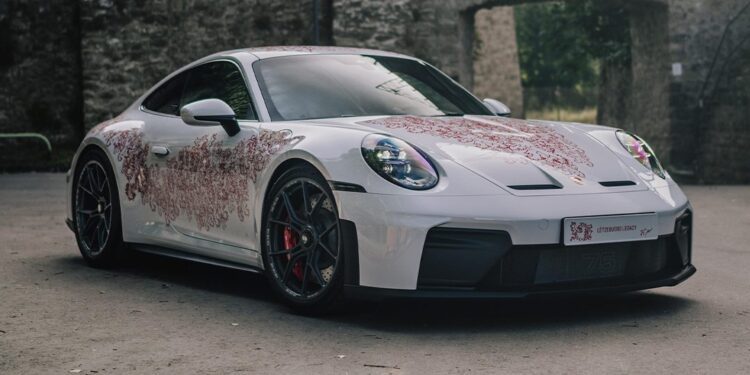 A Luxembourg-inspired Porsche Sonderwunsch 911 GT3 Touring with red floral patterns is parked on a paved surface in front of a stone wall with arched openings.