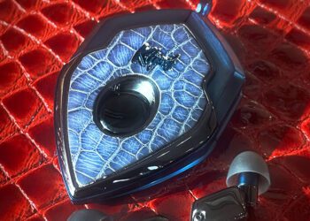 A metallic, shield-shaped electronic device with a blue textured surface sits on a red crocodile-patterned background, accompanied by two small black earphones—the perfect luxury accessories to upgrade your style.