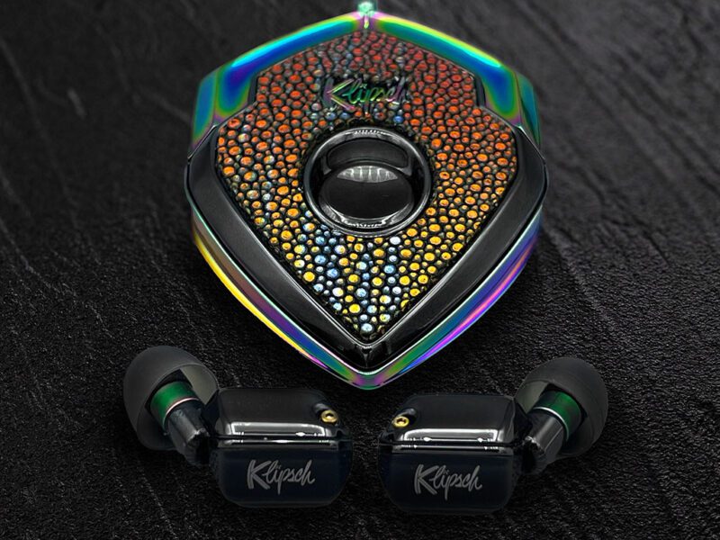 A pair of black Klipsch wireless earbuds with a jeweled, multi-colored triangular charging case on a dark textured surface—perfect for those seeking luxury accessories and a true style upgrade.
