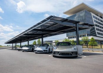 Four gray Porsche sports cars are parked under a large solar panel canopy at One Porsche Drive in Atlanta, beside a modern glass office building on a clear day.
