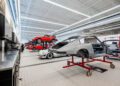 A spacious auto workshop with several classic cars, including a stripped-down Porsche body on a stand and red and white cars in various stages of repair, celebrates 10 years of automotive passion at One Porsche Drive Atlanta.