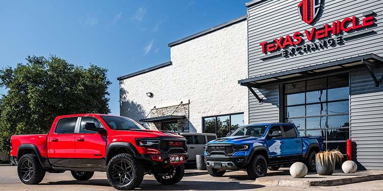 Two pickup trucks, one red and one blue, are parked in front of a building with a sign reading "Texas Vehicle Exchange," known for its selection of custom 4x4s and exotic cars, on a sunny day.