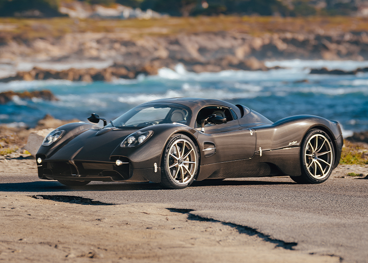 A sleek black roadster with aerodynamic design is parked on a coastal road, the ocean and rocky shoreline forming a stunning backdrop—reminiscent of the elegance seen at MIMO Milano Monza Motor Show.