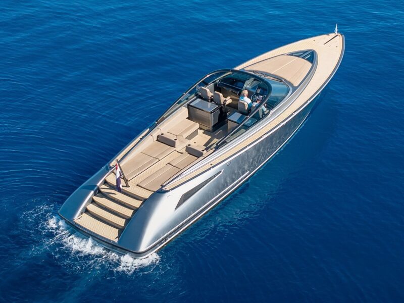 A modern, sleek motor yacht cruises on clear blue water, offering an unmatched maritime experience. Two people are seated at the helm with elegant wooden decking visible throughout this stunning example of luxury yachts.