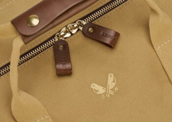 Close-up of a tan canvas bag with a gold metal zipper, brown leather zipper pulls, and a small gold bird logo with the letters "BW"—perfect for those looking to upgrade your style with luxury accessories.