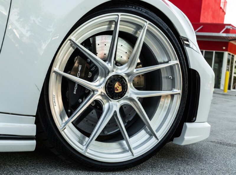 Close-up of ANRKY XR Wheels and brake disc on a white Porsche 911 Turbo S parked on a paved surface, with a red building in the background.