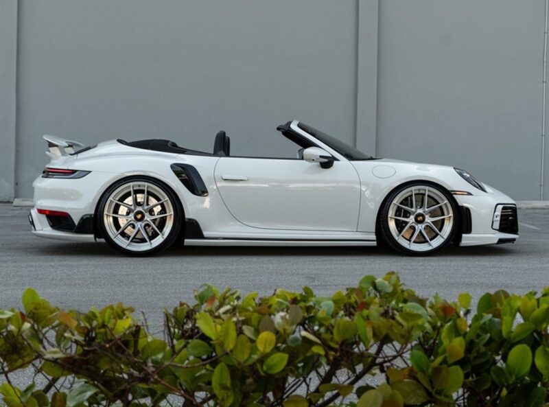 A white Porsche 911 Turbo S convertible with the top down is parked on pavement in front of a gray wall, with greenery in the foreground.