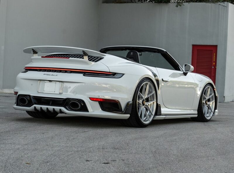 A white Porsche TechArt 911 Turbo S convertible with ANRKY XR Wheels is parked on a concrete surface near a grey building with a red door.
