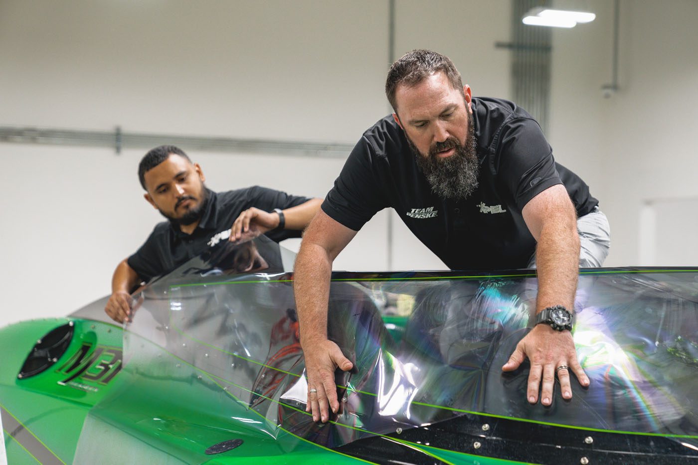 Two men in black shirts work together to fit a transparent Marine Protection Film canopy onto a bright green vehicle inside an indoor workshop.