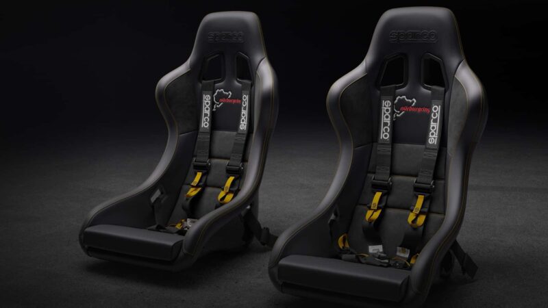 Two black Sparco racing seats with yellow seatbelt harnesses are positioned side by side on a dark surface, illuminated by soft lighting, evoking a performance-inspired look reminiscent of the Nürburgring.