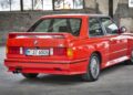 A red BMW E30 M3 coupe is parked indoors in an important industrial setting with a weathered concrete background.