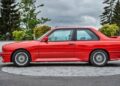 A red BMW E30 M3, an important classic sports car, is parked on a paved area, viewed from the side with trees and cloudy skies in the background.