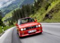 A red BMW E30 M3, an important icon among car enthusiasts, drives on a winding mountain road with trees and grassy hills in the background.