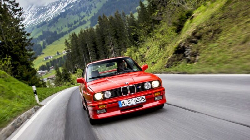 A red BMW E30 M3, an important icon among car enthusiasts, drives on a winding mountain road with trees and grassy hills in the background.