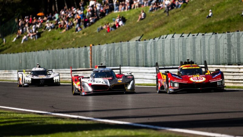 Three race cars, including a Hypercar Platform from top luxury brands, compete on a racetrack as spectators watch from a grassy hill in the background, evoking the spirit of Le Mans.