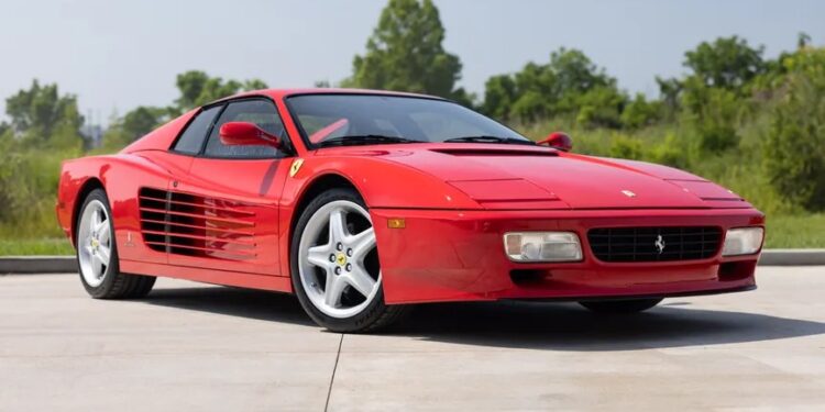 A rare Ferrari 512 TR Prototype sports car is parked on a driveway with greenery and trees in the background under a clear sky.