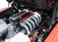 A close-up view of a 1992 Ferrari 512 TR Prototype engine with red valve covers and multiple intake manifolds, housed within a red pre-production vehicle with the hood open.