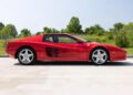 A rare Ferrari Testarossa sports car is parked on a paved surface with grass and trees in the background under a clear sky.