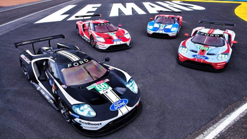 Four Ford GT race cars are parked on the Le Mans racetrack near large white "LE MANS" letters painted on the asphalt, capturing the spirit of Ford Le Mans and evoking memories of the legendary Ford vs Ferrari rivalry.