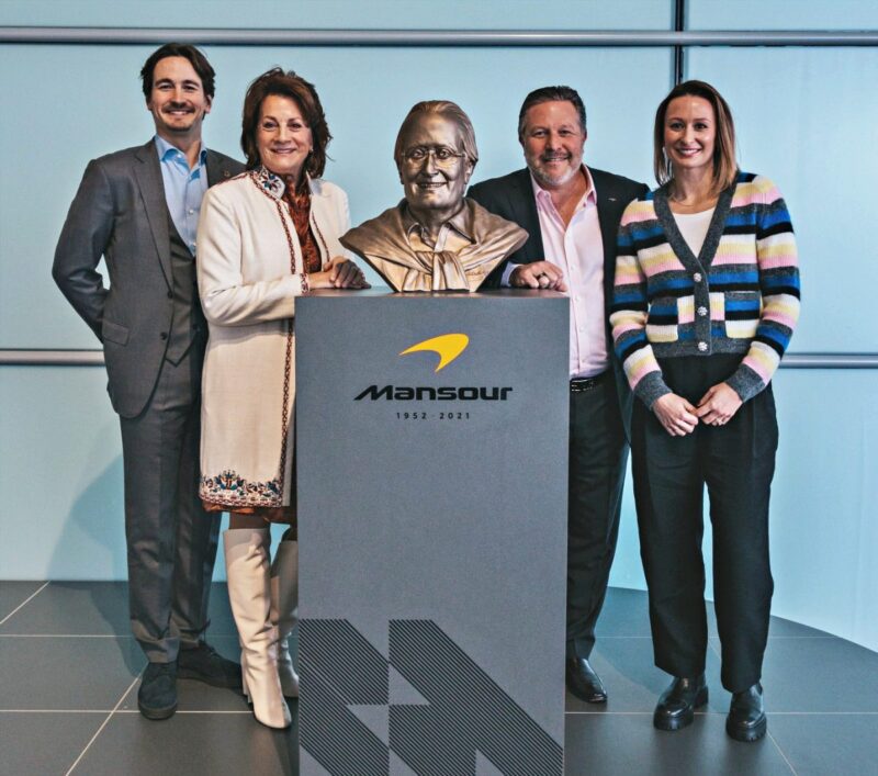 Four people stand beside a bronze bust on a podium labeled "Mansour 1932-2021" in a modern indoor setting, echoing the exclusivity of the 20 Ultra-Exclusive 1-of-1 McLarens being offered in a historic sale.