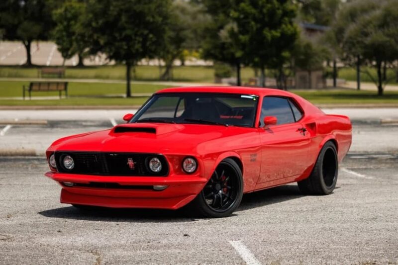 A red classic muscle car with black wheels is parked in an empty lot, surrounded by grass and trees—a timeless icon for fans of high-performance cars.