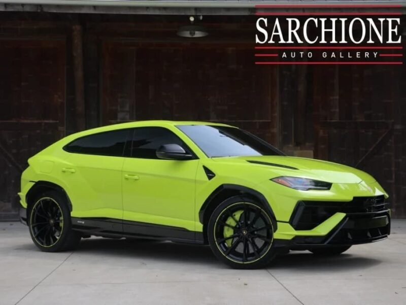A bright green Lamborghini Urus in stunning Verde Scandal is parked in front of a dark wooden building, with “Sarchione Auto Gallery” signage in the top right corner.