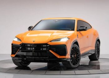 An orange Lamborghini Urus luxury car is displayed in a well-lit studio with a plain background, showing a front and side view of the vehicle.