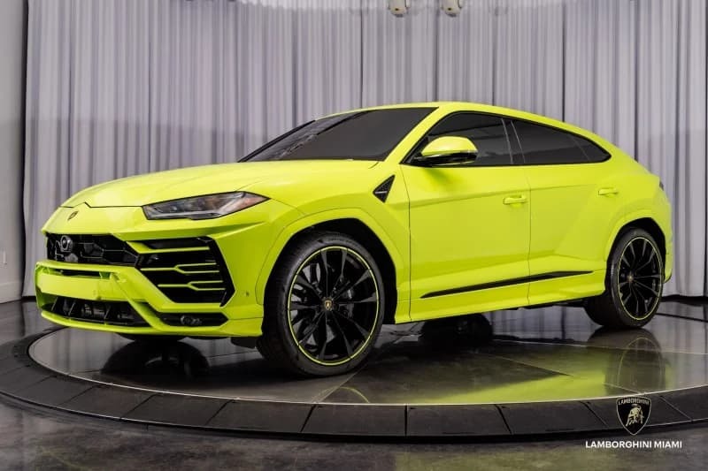 A neon yellow Lamborghini Urus SUV, finished in striking Verde Scandal, is displayed indoors on a rotating platform with a white curtain backdrop, telling an unforgettable Lamborghini story.