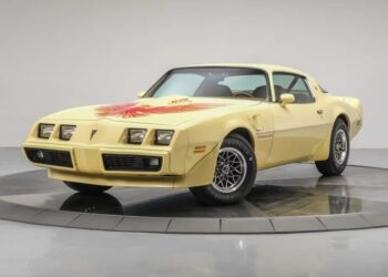 A yellow 1979 Pontiac Firebird Trans Am with a hood decal is parked on a rotating display platform in a white studio, showcasing one of the iconic Classic Muscle Cars enthusiasts dream of.