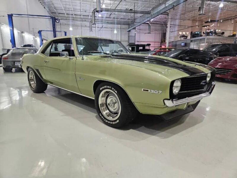 A classic light green Chevrolet Camaro 307 with black racing stripes is parked inside a spacious, well-lit garage with other Classic Muscle Cars in the background.