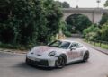 A white Porsche Sonderwunsch sports car with red detailing drives on a curving road surrounded by greenery and a stone bridge, evoking a Luxembourg-inspired scene.