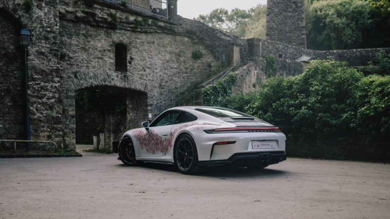 A white 911 GT3 Touring with red decorative patterns, inspired by Porsche Sonderwunsch, is parked in front of old stone walls surrounded by lush greenery.