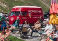 People sit on outdoor chairs and loungers near a red vintage Porsche-branded van, with colorful umbrellas and mountains in the background, capturing a relaxed FAT Mankei vibe.