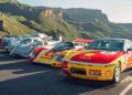 A row of classic Porsche race cars, including a Shell-liveried model and a FAT Mankei 963 RSP, parked on a mountain road with green hills and rocky cliffs in the background.