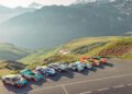 A row of classic and modern sports cars, including a Porsche 963 RSP by FAT Mankei, is parked on a mountain road, overlooking a scenic valley and snow-capped peaks.