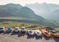 Seven Porsche race cars, including the 963 RSP by FAT Mankei, are parked in a row on a mountain overlook with green hills and distant peaks in the background.