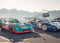 Two Porsche race cars, including the iconic FAT Mankei 963 RSP, parked side by side on a mountain road with distant peaks and a partly cloudy sky in the background.