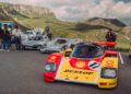 Several vintage race cars, including a Porsche and a Shell-Dunlop 963 RSP, are parked on a mountain road with people gathered nearby and green hills in the background near FAT Mankei.