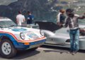 A person in racing gear poses for a selfie next to a vintage Porsche rally car and a 963 RSP race car, with several people and mountains in the background.