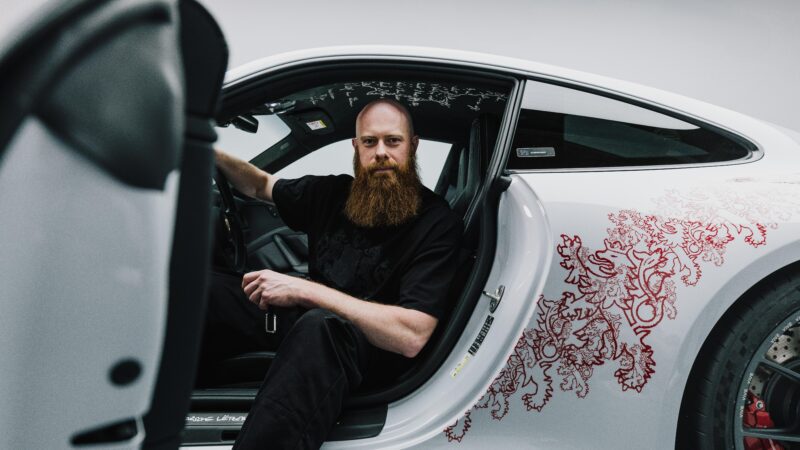 A man with a long beard sits in the driver's seat of a white Porsche Sonderwunsch 911 GT3 Touring, featuring Luxembourg-inspired red decorative designs on the door and body.