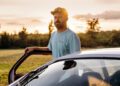 A man wearing a cap and light blue t-shirt stands beside the open door of a Porsche in a grassy field at sunset, hinting at an origin story set against rarest colors of the golden hour.