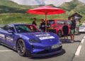 A blue Porsche Taycan with event stickers is parked roadside near a red umbrella, with several people, other Porsche cars, and a FAT Mankei 963 RSP in the background, set against a mountainous backdrop.