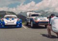 A person in a Porsche shirt photographs two vintage Porsche rally cars, including the 963 RSP, parked outdoors with mountains in the background.