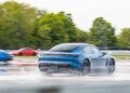 A blue Porsche speeds through water on a wet race track at the Toronto Experience Center, with two other cars in the background and trees lining the track.