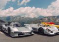 Three classic Porsche race cars, including the 963 RSP and a FAT Mankei-liveried model, are parked on a mountain road with green peaks and a partly cloudy sky in the background.